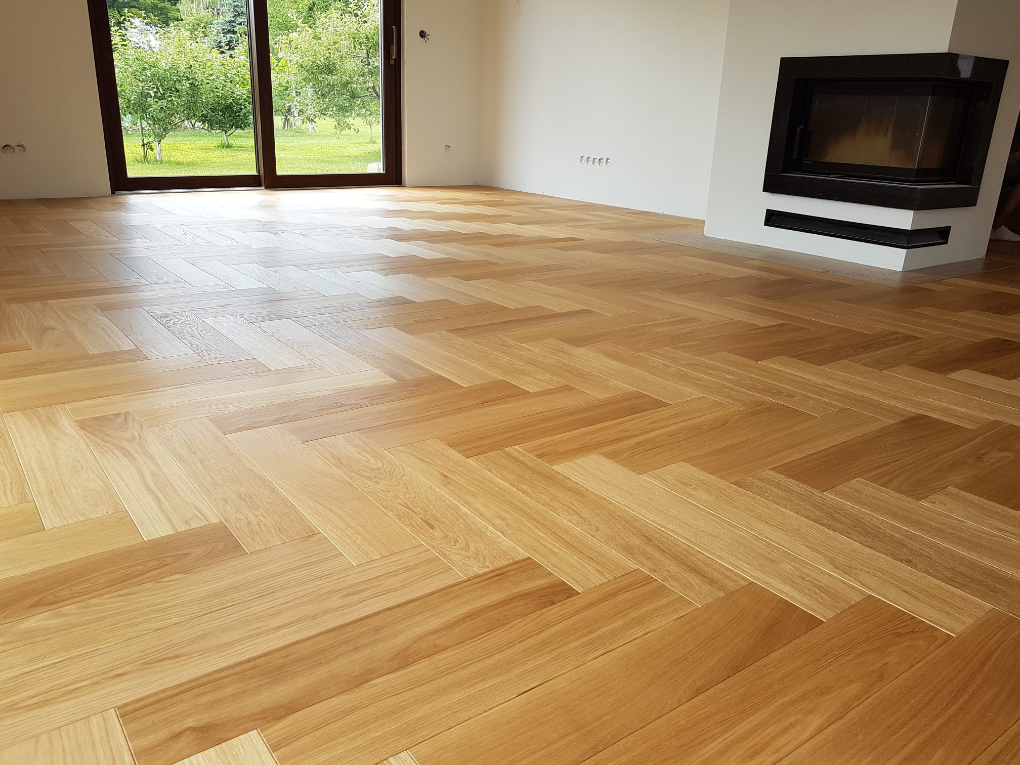 Jasny parkiet ułożony w jodełkę francuską w przestronnym salonie z kominkiem i widokiem na ogród przez przesuwne okno.