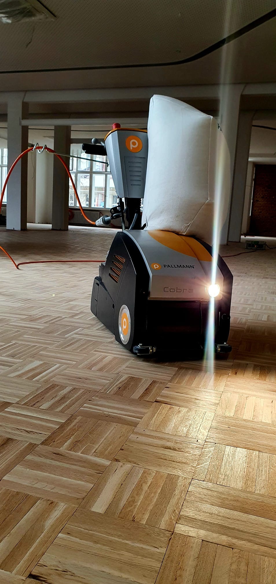 Maszyna Pallmann Cobra do renowacji parkietu w trakcie pracy, widoczny kabel zasilający i worek na pył, parkiet ułożony w kwadraty, surowe wnętrze budynku.