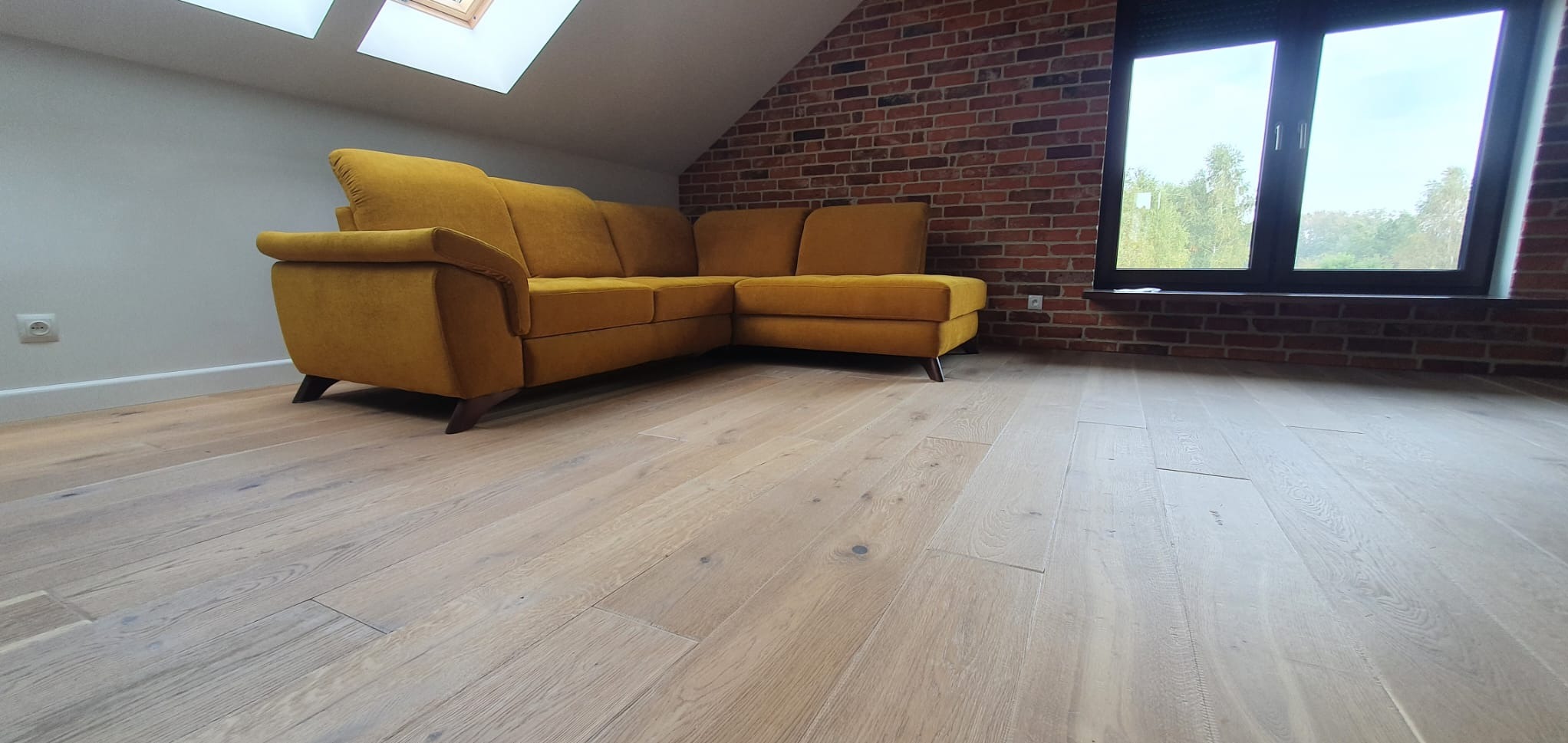 Jasne, drewniane deski podłogowe w pomieszczeniu w Żarach z żółtą sofą narożną i ścianą z czerwonej cegły, widok z niskiego kąta.
