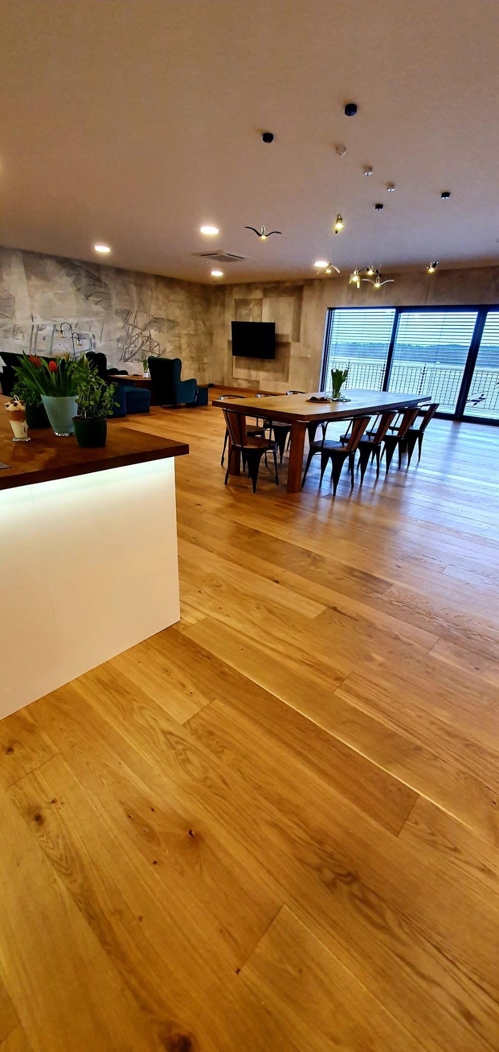 Salon z jasną, drewnianą podłogą ułożoną w jodełkę, widok na stół z krzesłami, kanapę i okno z widokiem na zewnątrz.