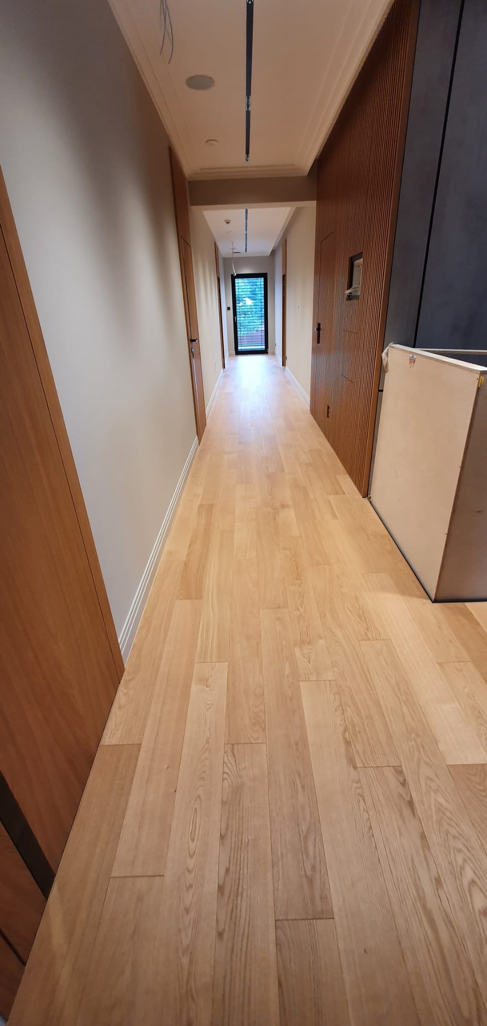 Jasna, drewniana podłoga w korytarzu z widocznymi detalami usłojenia, prowadząca do przeszklonych drzwi na końcu, z drewnianymi panelami ściennymi po prawej stronie i gładkimi ścianami po lewej.