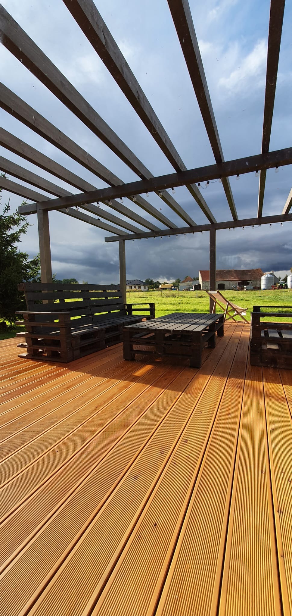 Rustykalny taras z desek kompozytowych w odcieniach miodowego brązu, z meblami z palet pod drewnianą pergolą, w tle zielone pole i zachmurzone niebo.