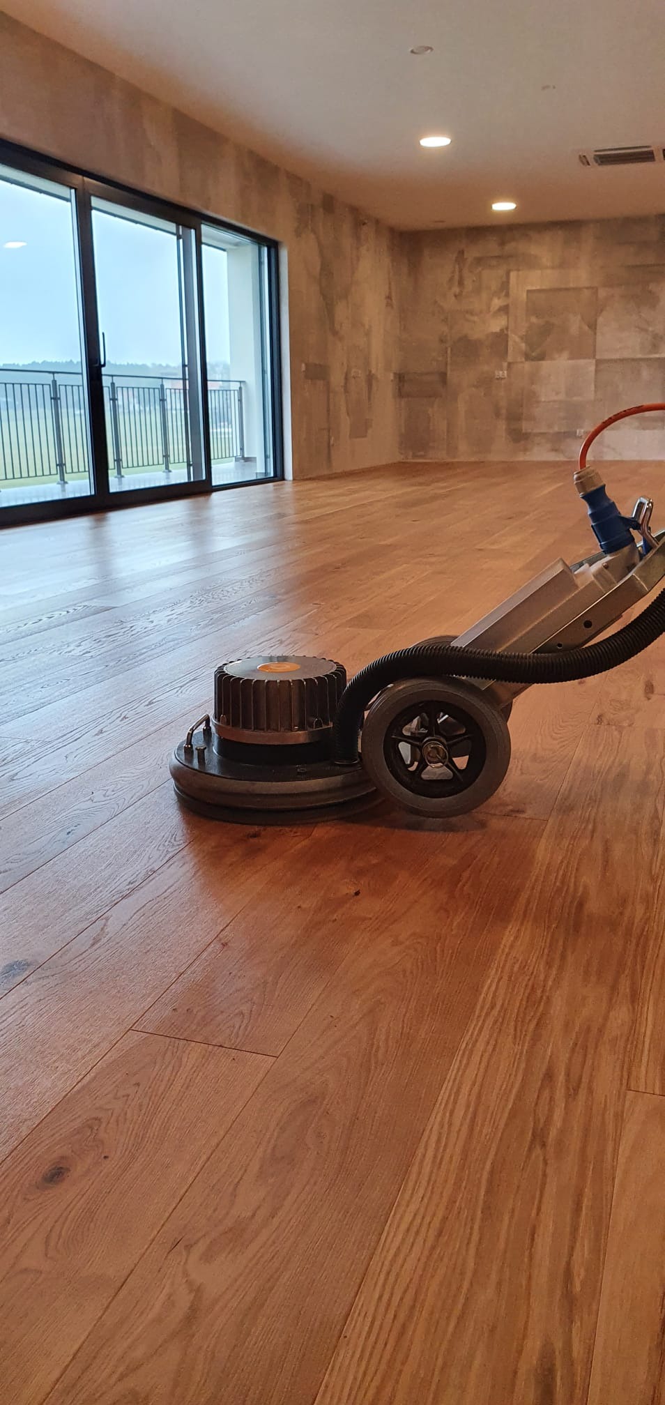 Maszyna do renowacji parkietu w trakcie pracy, widoczne świeżo odnowione deski, duże okno balkonowe w tle.