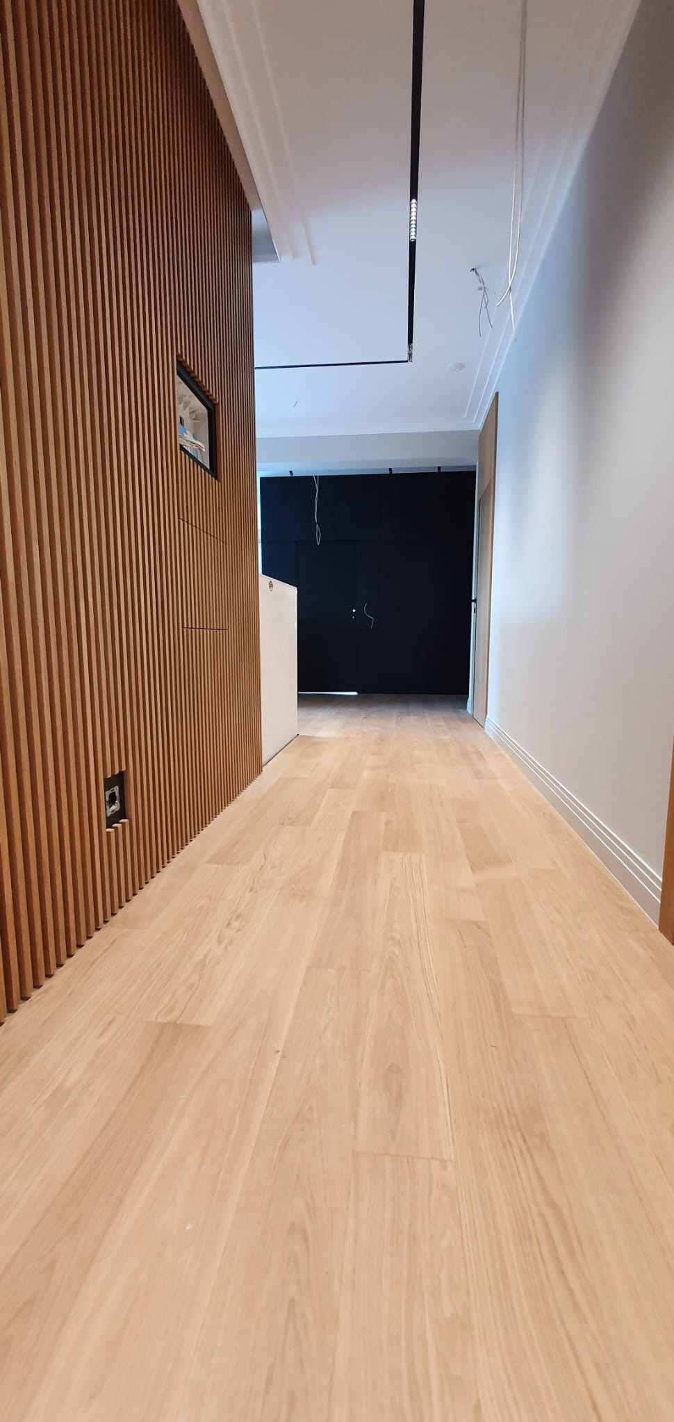 Jasna, drewniana podłoga w korytarzu, z widoczną ścianą z drewnianych listew i czarną zabudową w tle, oświetlona nowoczesną lampą sufitową.
