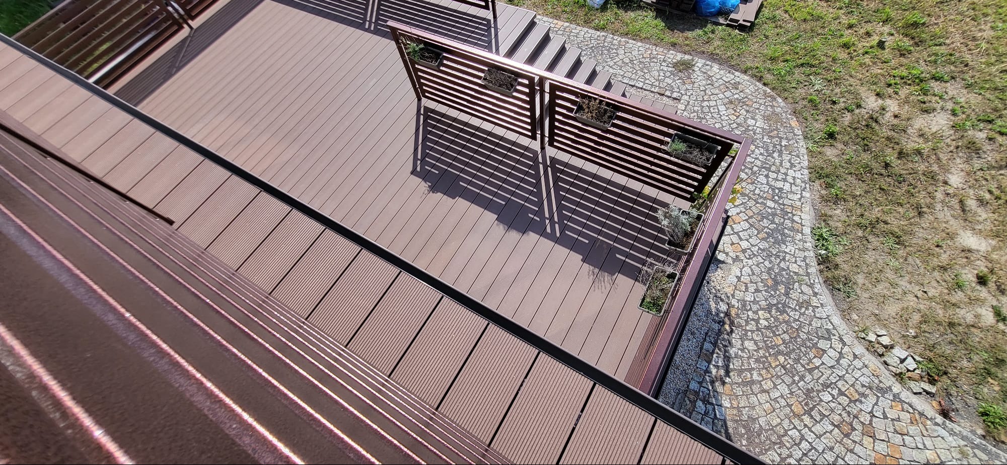 Widok z góry na brązowy taras kompozytowy z drewnianą balustradą z donicami, obok ścieżka z kostki brukowej i trawnik.