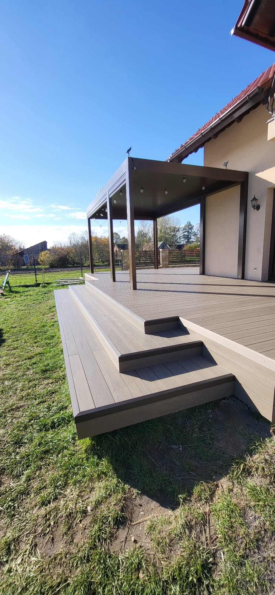 Nowoczesny taras kompozytowy z zadaszeniem w kolorze brązowym, schody wejściowe, widok na dom i ogród w słoneczny dzień.