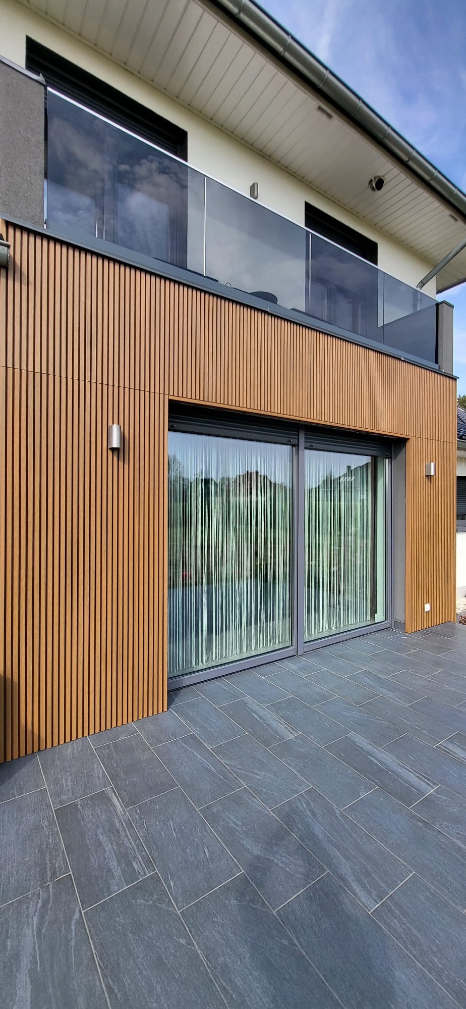 Nowoczesna elewacja budynku z drewnopodobnymi panelami pionowymi, dużymi przesuwnymi oknami z widokiem na ogród i tarasem wyłożonym szarymi płytami.
