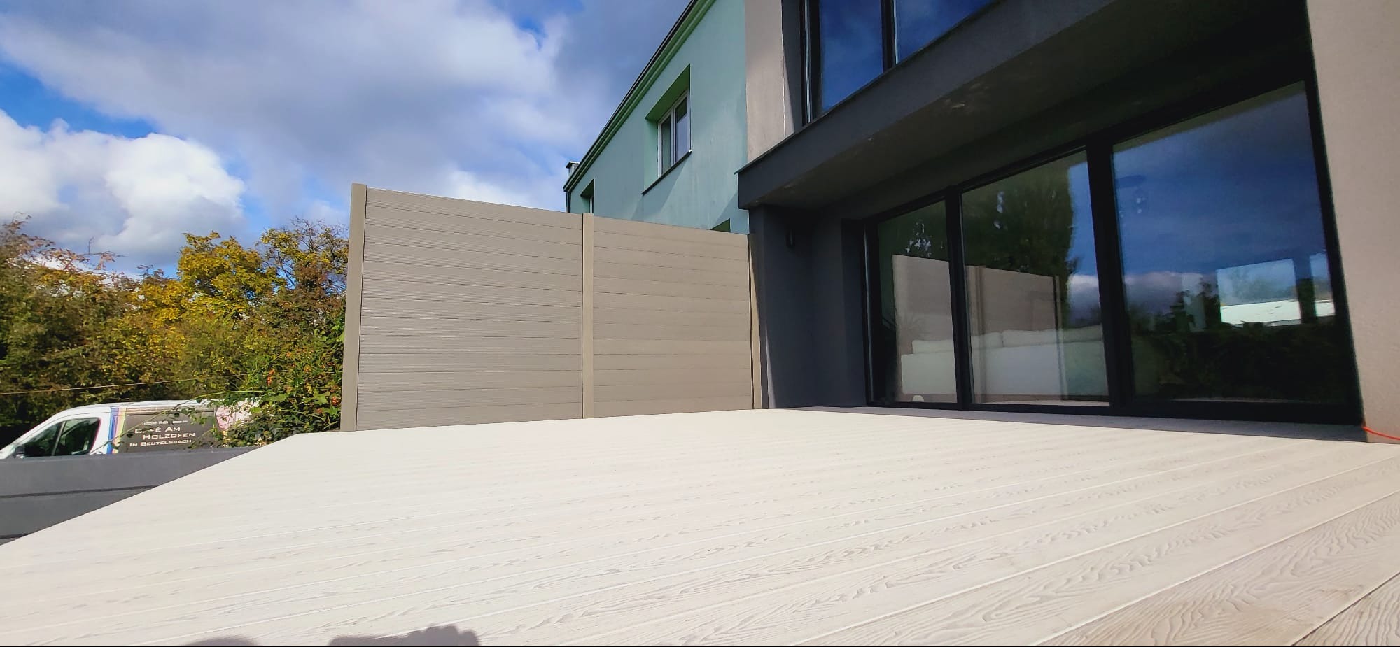 Jasny, kompozytowy taras z widoczną strukturą drewna, przylegający do nowoczesnego domu z dużymi oknami przesuwnymi i panelową ścianą, widoczny fragment firmowego samochodu w tle.