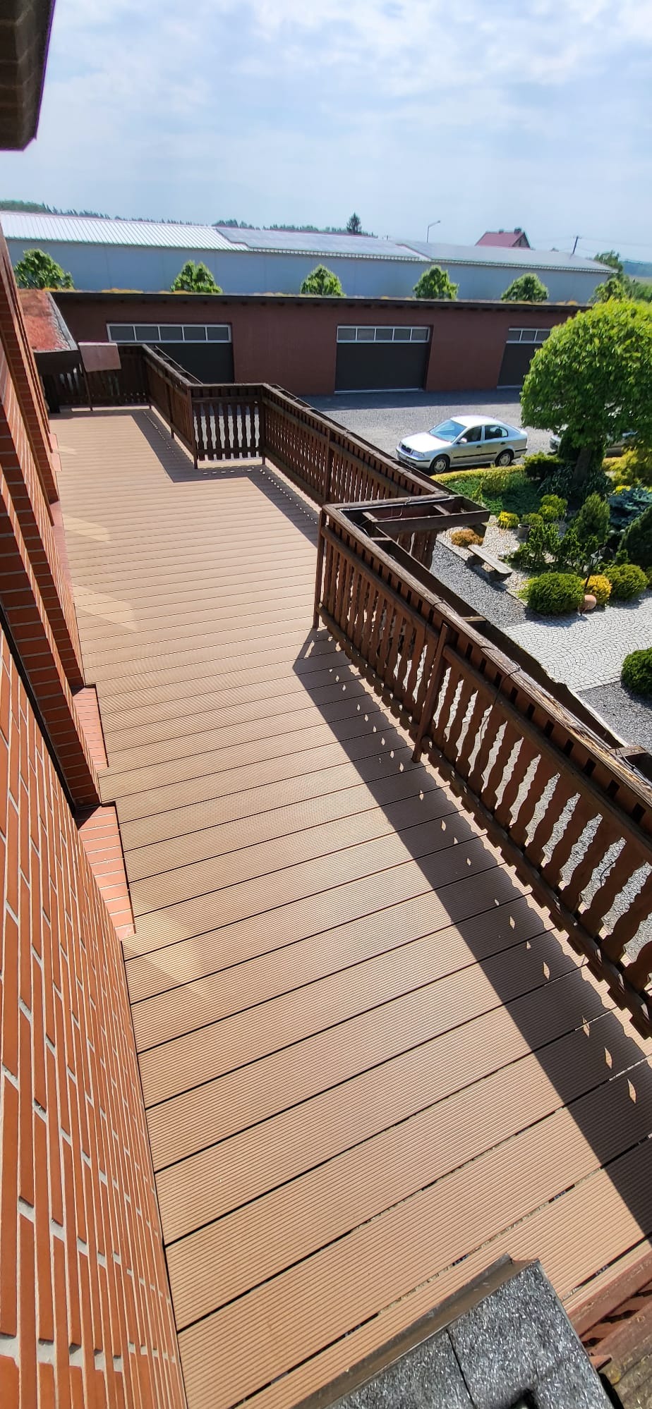 Jasny, kompozytowy taras z drewnianą balustradą, widok z góry na ogród z ozdobnymi krzewami i zaparkowany samochód.