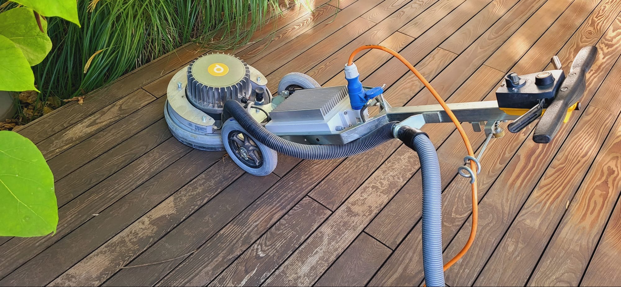 Szlifowanie drewnianego tarasu przy użyciu profesjonalnej szlifierki tarczowej z odkurzaczem, widoczne ślady po szlifowaniu i fragment roślinności w tle.