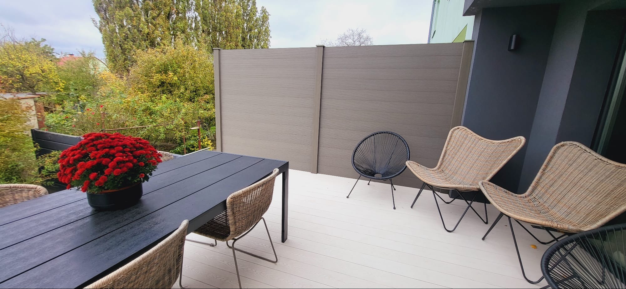 Nowoczesny taras z jasną kompozytową podłogą, otoczony panelami, z czarnym stołem, wiklinowymi krzesłami i czerwonymi kwiatami w donicy.