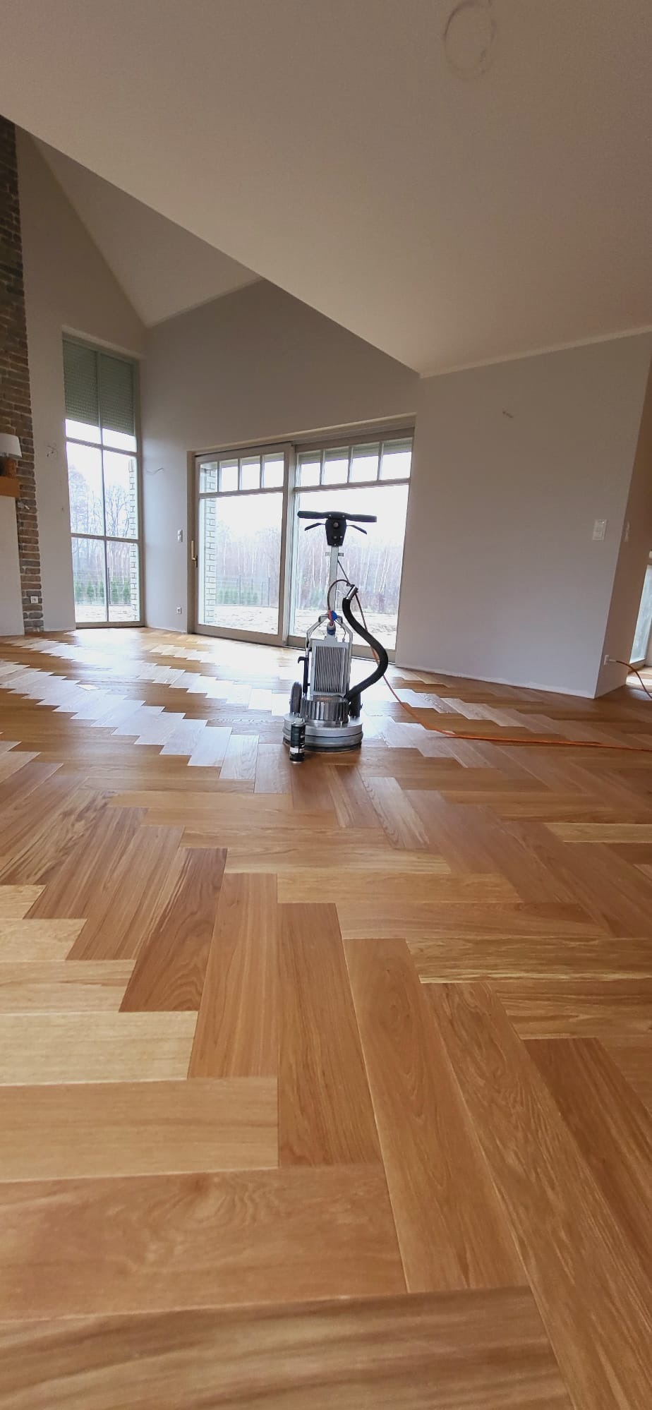 Parkiet ułożony w jodełkę podczas cyklinowania maszyną z podłączonym odkurzaczem, widoczne duże okna i fragment ściany z cegły.