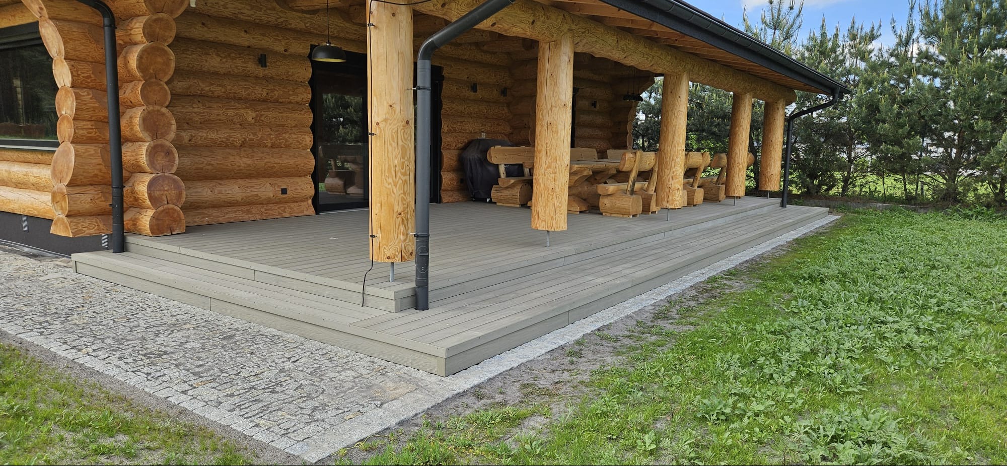Zadaszony taras z kompozytowych desek w odcieniach szarości, przylegający do drewnianego domu z bali, z widocznym drewnianym stołem i ławkami oraz kamienną ścieżką.