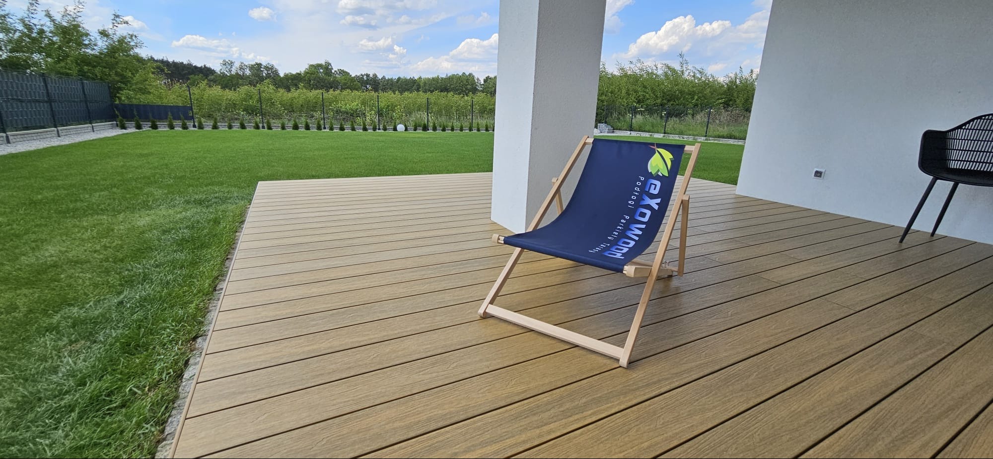 Nowoczesny taras z desek kompozytowych w odcieniach brązu, z leżakiem reklamowym i czarnym krzesłem, widok na zadbany trawnik i zieleń.