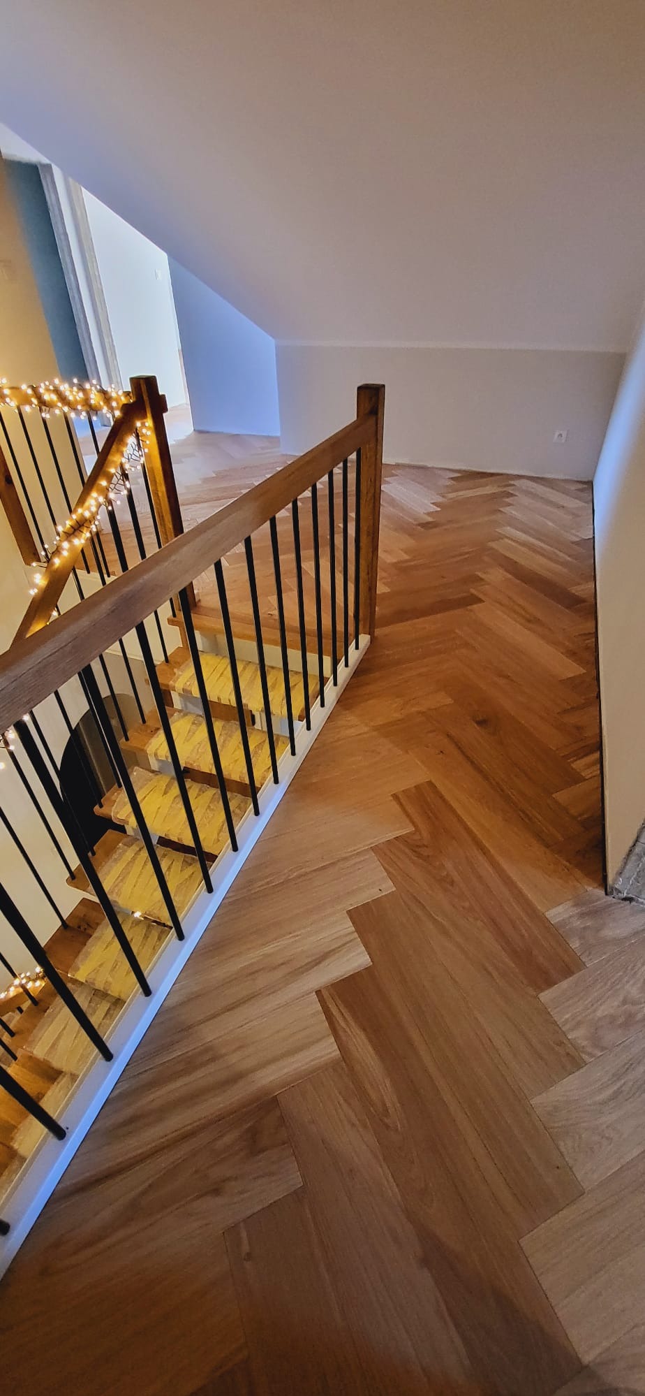 Parkiet ułożony w jodełkę na podłodze i schodach, drewniane balustrady z czarnymi, metalowymi prętami ozdobione lampkami.