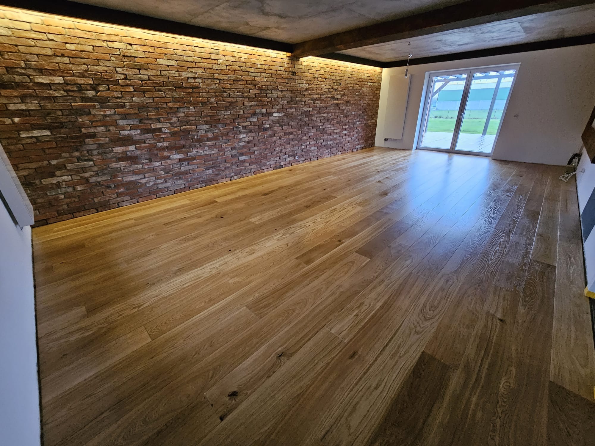 Jasna, lakierowana podłoga z desek ułożona w przestronnym, surowym wnętrzu z ceglaną ścianą i dużym oknem tarasowym.