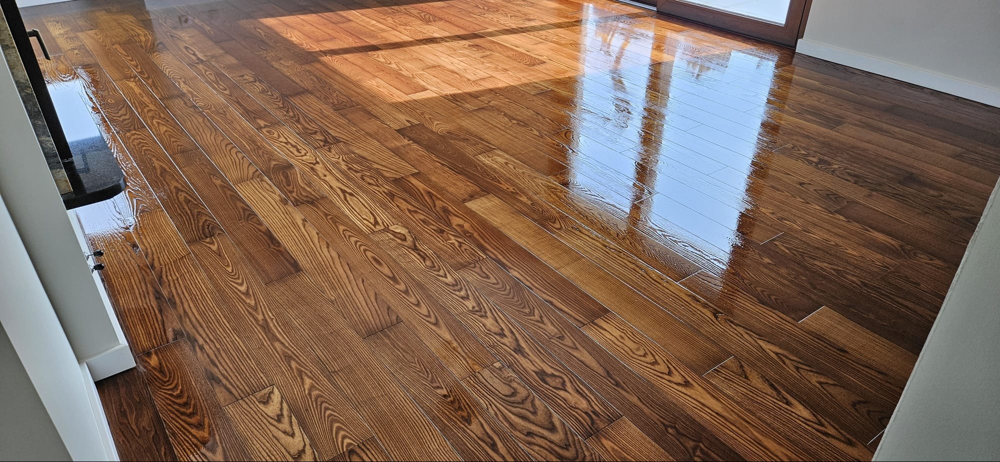 Błyszcząca, świeżo polakierowana drewniana podłoga z wyraźnym usłojeniem, odbijająca światło słoneczne wpadające przez okno balkonowe.