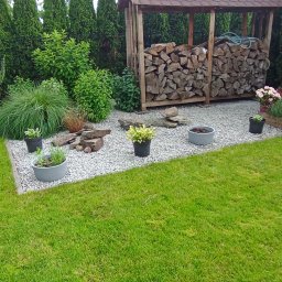 Ogród Dom - Uporządkowana przestrzeń ogrodu z równo przystrzyżonym trawnikiem, wyłożona białym kamieniem, z drewnianą wiatą na drewno, ozdobnymi kamieniami i donicami z roślinami.