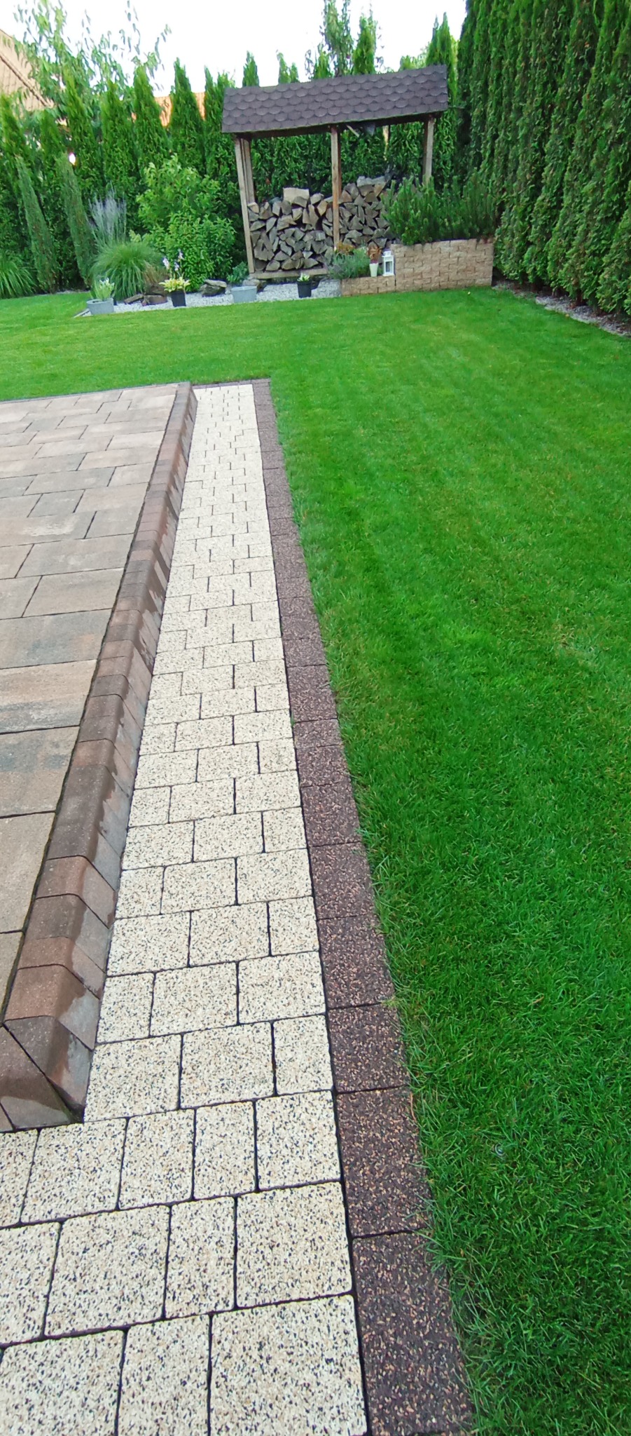 Ułożony chodnik z kostki brukowej w ogrodzie, z obrzeżem z ciemniejszej kostki, przylegający do zadbanego trawnika; w tle drewniana wiata na drewno i różnorodna roślinność.