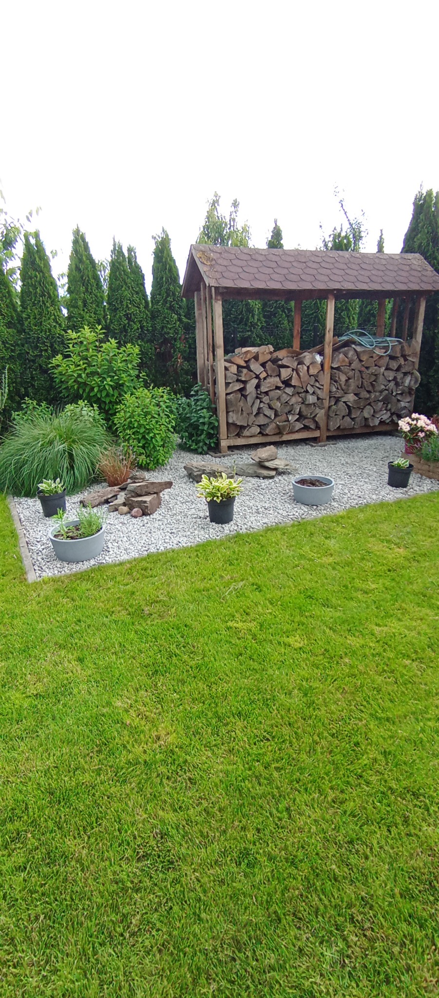Uporządkowana przestrzeń ogrodu z równo przystrzyżonym trawnikiem, wyłożona białym kamieniem, z drewnianą wiatą na drewno, ozdobnymi kamieniami i donicami z roślinami.