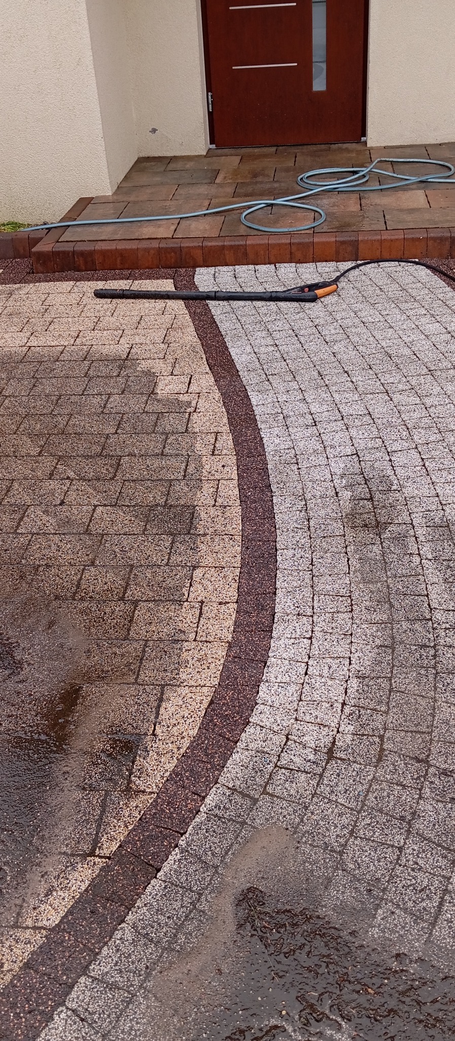 Czyszczenie kostki brukowej przed domem za pomocą myjki ciśnieniowej, widoczny efekt przed i po, z ciemniejszą, mokrą powierzchnią po lewej i jaśniejszą, czystą po prawej stronie. W tle wejście...