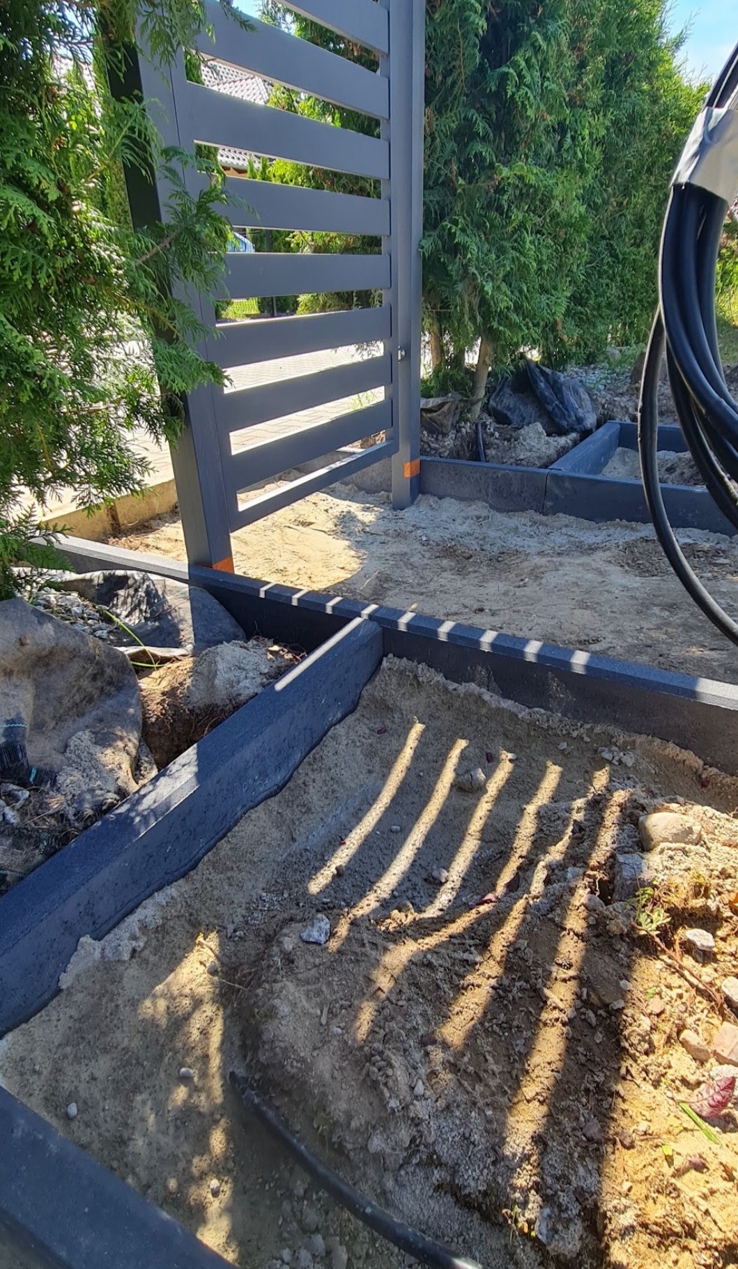 Montaż nowoczesnego ogrodzenia panelowego w kolorze grafitowym, widoczne obrzeża rabat w trakcie tworzenia, piasek i cień rzucany przez ogrodzenie.