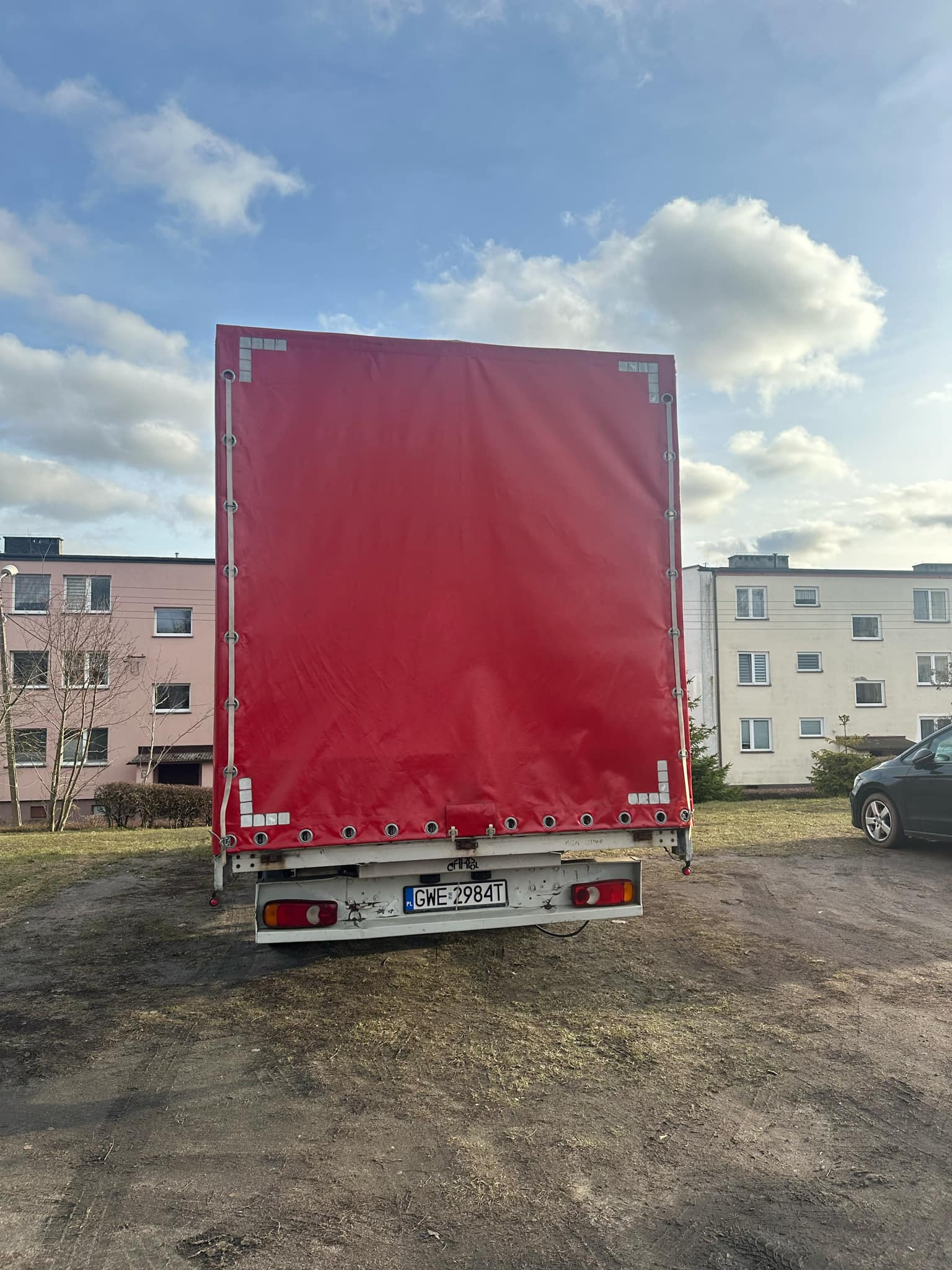 Tył czerwonej naczepy ciężarówki z widoczną tablicą rejestracyjną GWE 2984T na tle osiedla mieszkaniowego i pochmurnego nieba.