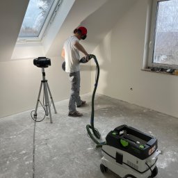 Mirosław Nieśpiał Mr Handy - Pracownik w czerwonej czapce szlifuje ścianę na poddaszu za pomocą szlifierki Festool z odkurzaczem przemysłowym, widoczne okno dachowe i okno pionowe.
