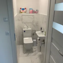 Mała, odnowiona toaleta z marmurowymi płytkami, wiszącą umywalką i podwieszanym WC; widoczny fragment białych drzwi z przeszkleniem.