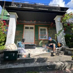 Mirosław Nieśpiał Mr Handy - Przygotowania do malowania elewacji domu z zielonymi ścianami i białymi kolumnami, okna i drzwi zabezpieczone folią, pracownik siedzi na schodach.