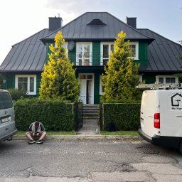 Mirosław Nieśpiał Mr Handy - Zielony dom z ciemnym dachem po remoncie, z zaparkowanymi samochodami i pracownikiem siedzącym na chodniku przed posesją.