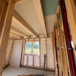 Wnętrze w trakcie budowy, widoczne drewniane ramy ścian, izolacja z wełny mineralnej, częściowo zamontowane płyty gipsowo-kartonowe na suficie, instalacja elektryczna i rury grzewcze w ścianach.