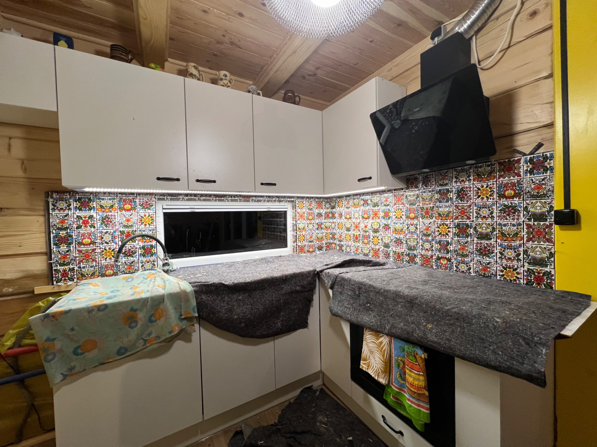 Fragment kuchni w drewnianym domu z białymi szafkami i wzorzystą mozaiką na ścianie, blat zabezpieczony szarą włókniną, widoczny zlew przykryty tkaniną w kwiaty.