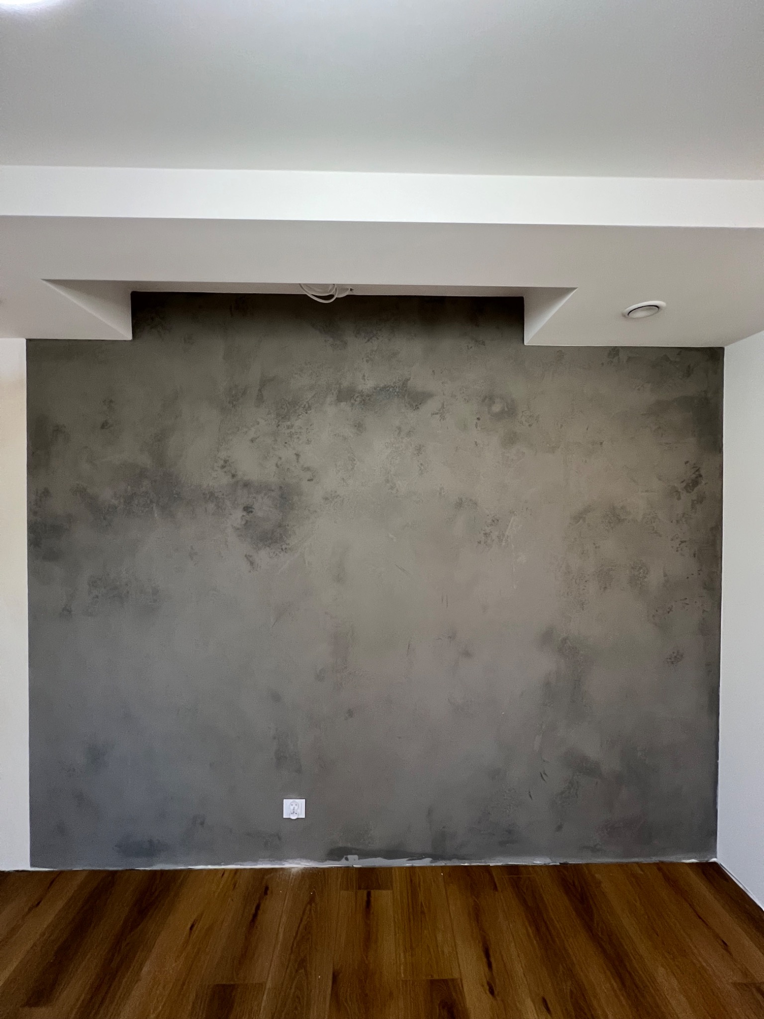 Ściana z dekoracyjną szarą fakturą, widoczne drewniane panele podłogowe i białe wykończenia sufitu z wnęką na oświetlenie.