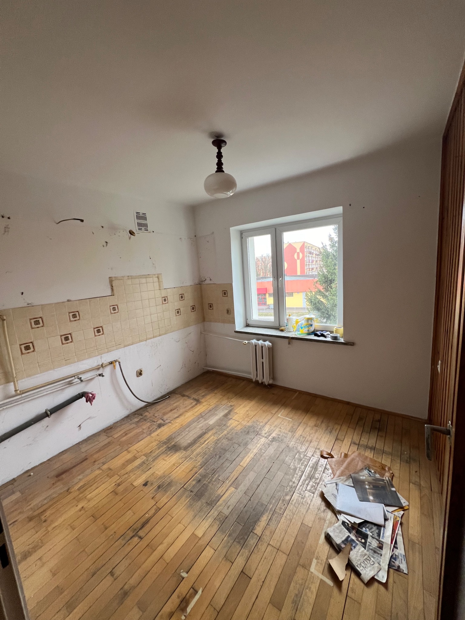 Kuchnia przed remontem: fragment ściany z częściowo usuniętymi płytkami, odsłonięte rury, drewniana podłoga z widocznymi śladami użytkowania i sterta gazet w rogu.