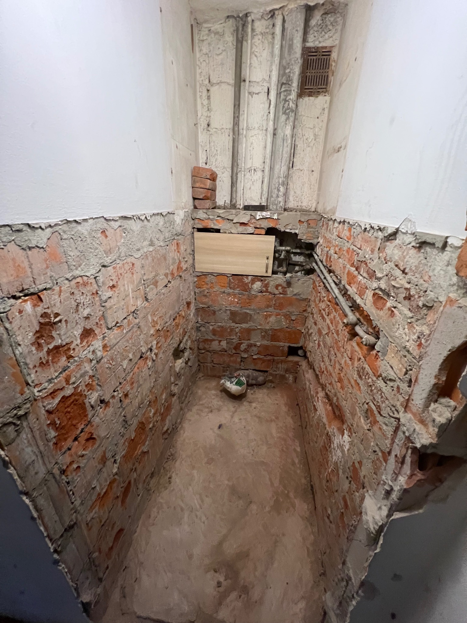 Niedokończona wnęka w łazience z odsłoniętą czerwoną cegłą, widoczne rury, fragment drewnianej płyty i biała ściana w górnej części.