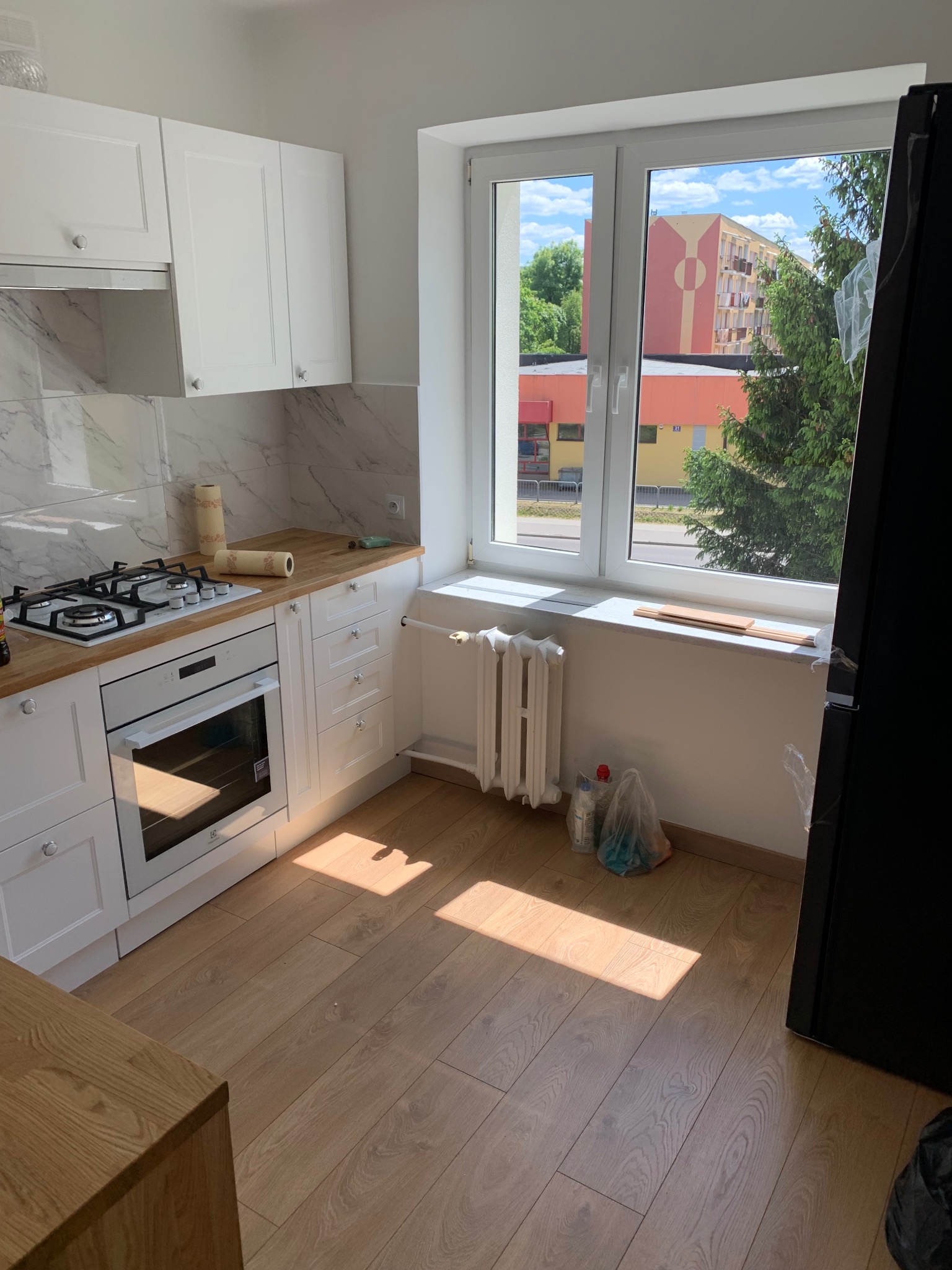 Jasna kuchnia z białymi szafkami, drewnianym blatem i panelami podłogowymi, widok na budynek przez okno.