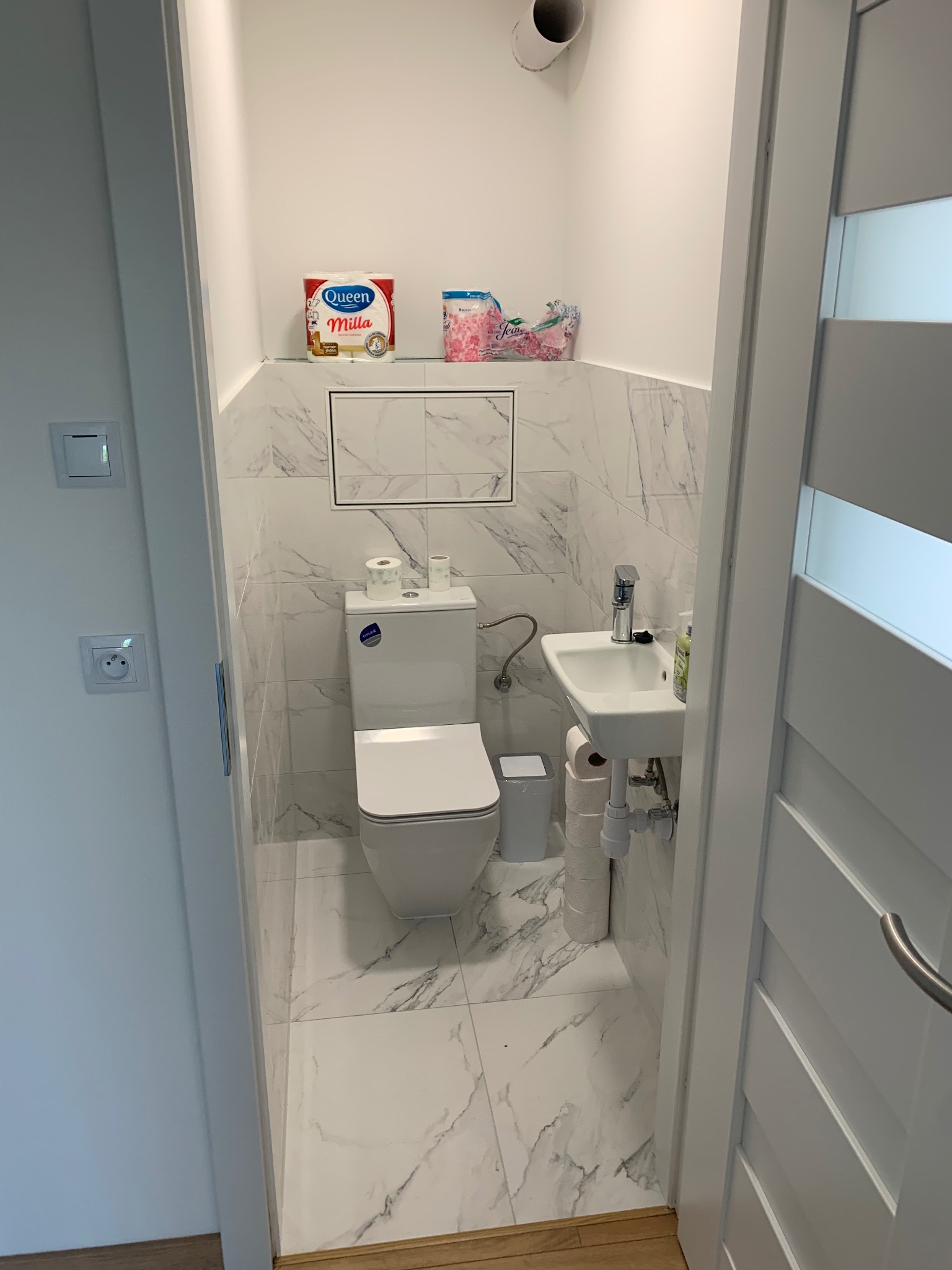 Mała, odnowiona toaleta z marmurowymi płytkami, wiszącą umywalką i podwieszanym WC; widoczny fragment białych drzwi z przeszkleniem.