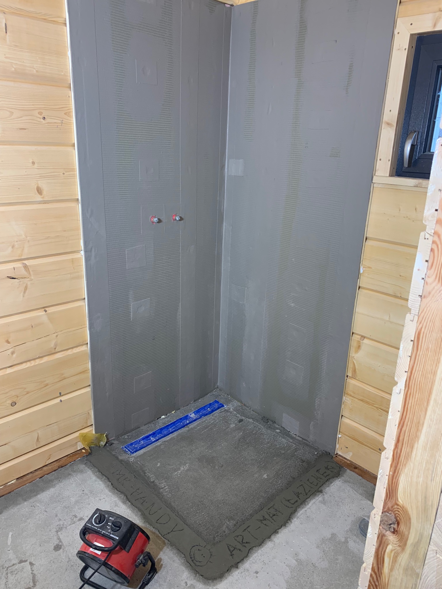 Przygotowana do wyłożenia płytkami kabina prysznicowa w drewnianym domku, ściany pokryte szarą warstwą izolacyjną, na podłodze wylewka z napisem 'MR. VANDY ART-MAT (ŁAZIENEK)', obok grzejnik.