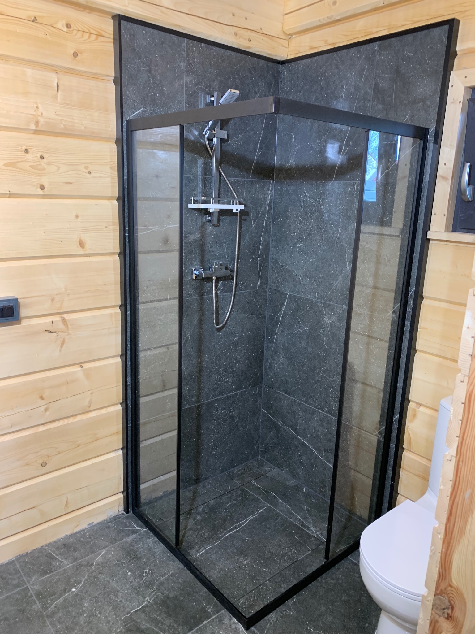 Nowoczesna kabina prysznicowa z czarną ramą na tle ściany z płytek imitujących marmur w łazience wykończonej drewnem.