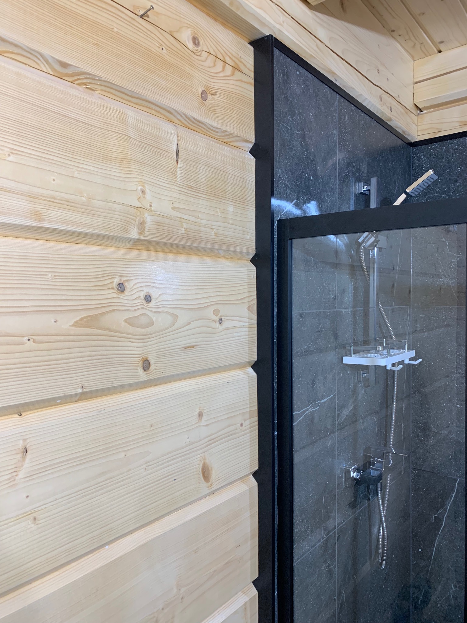 Fragment łazienki w domu z bali: kabina prysznicowa z czarną ramą i marmurowymi płytkami, sąsiadująca z jasną, drewnianą ścianą z widocznymi słojami.