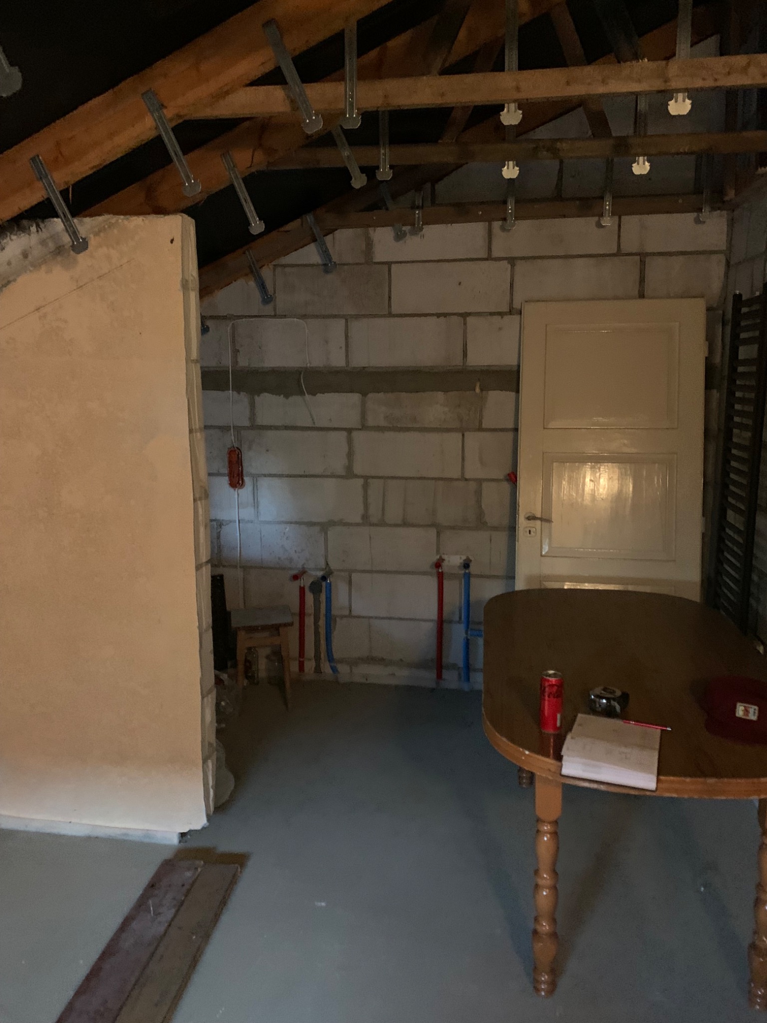 Poddasze w trakcie remontu: widoczne surowe ściany z bloczków, niezabudowane belki stropowe z metalowymi elementami, częściowo otynkowana ściana, rury instalacyjne i stolik z notatkami, puszką...