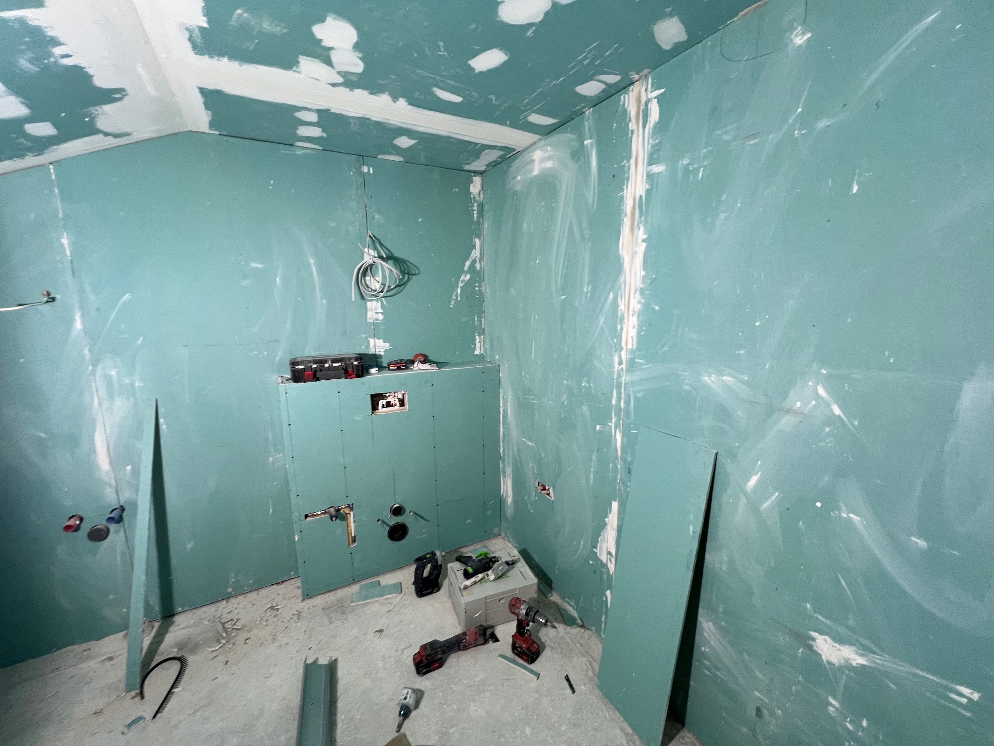 Wnętrze łazienki w trakcie prac wykończeniowych, widoczne płyty kartonowo-gipsowe na ścianach z widocznymi śladami szpachlowania, przygotowane podłoże pod armaturę oraz narzędzia budowlane...