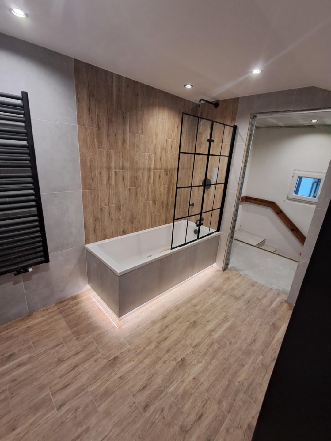 Nowoczesna łazienka z wanną obudowaną szarymi płytkami, czarną kabiną prysznicową typu walk-in i podświetleniem LED u dołu wanny oraz grzejnikiem drabinkowym.