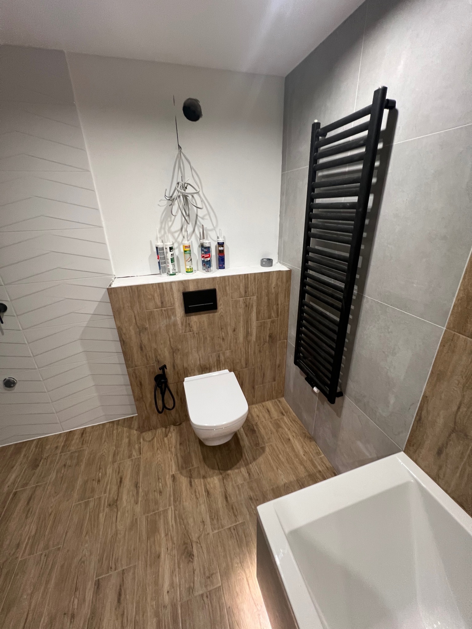 Wnętrze łazienki w trakcie prac wykończeniowych: widoczna biała toaleta podwieszana, grzejnik drabinkowy w kolorze czarnym, biała wanna, częściowo ułożone płytki imitujące drewno na podłodze...