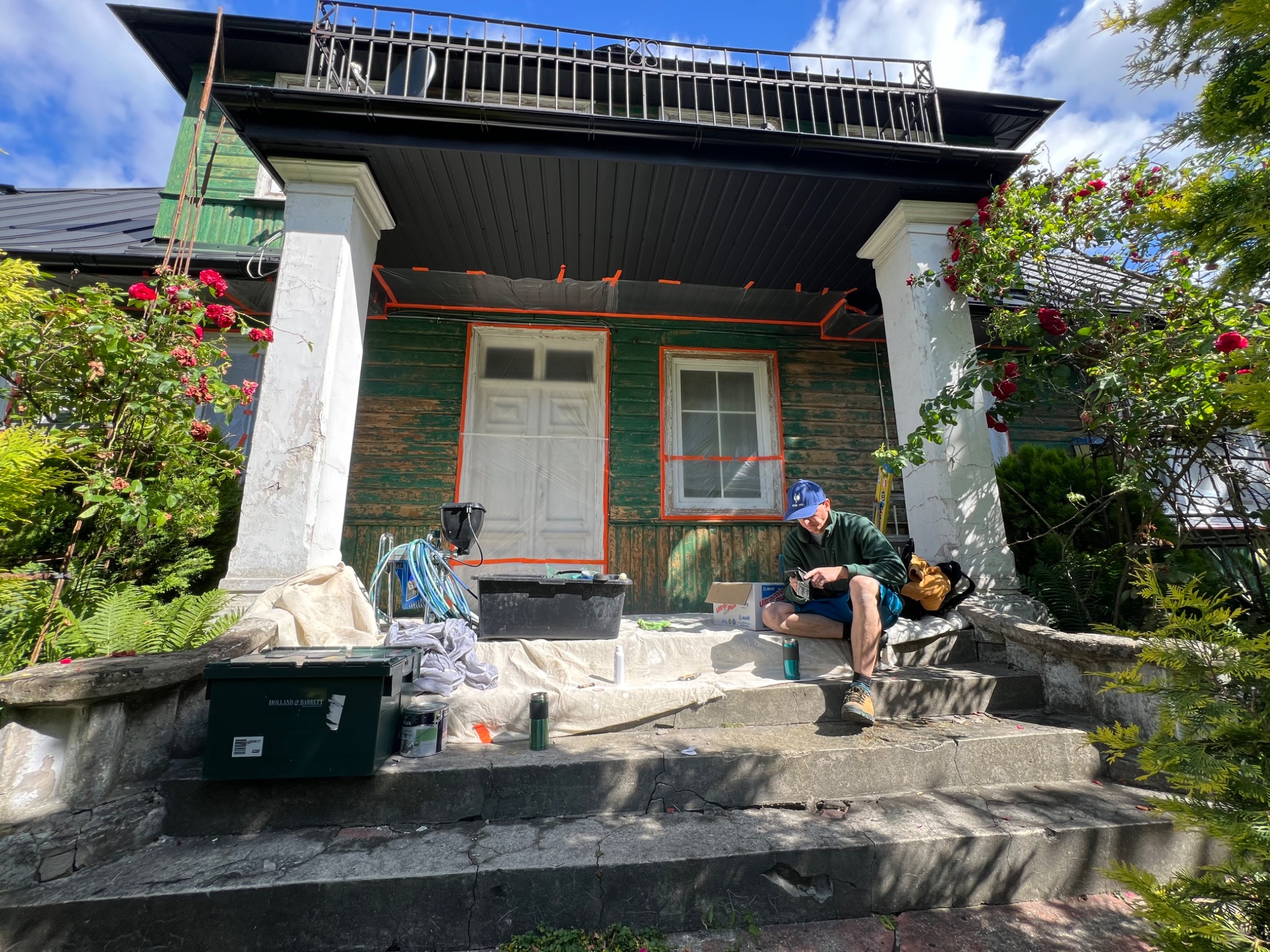 Przygotowania do malowania elewacji domu z zielonymi ścianami i białymi kolumnami, okna i drzwi zabezpieczone folią, pracownik siedzi na schodach.