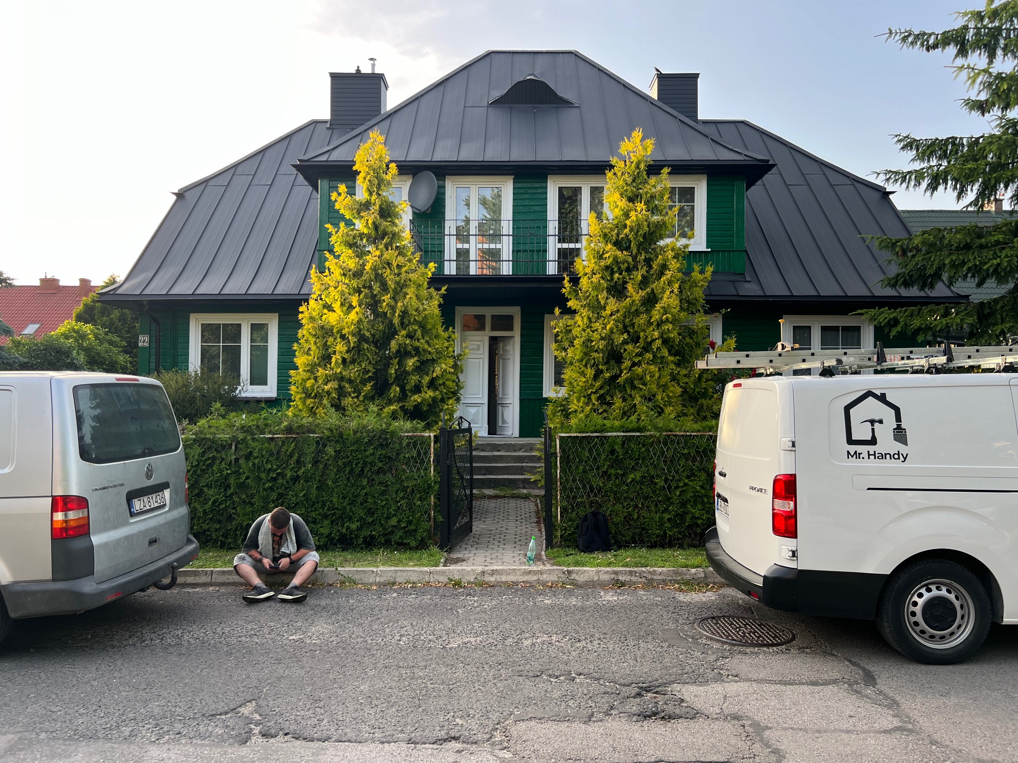 Zielony dom z ciemnym dachem po remoncie, z zaparkowanymi samochodami i pracownikiem siedzącym na chodniku przed posesją.