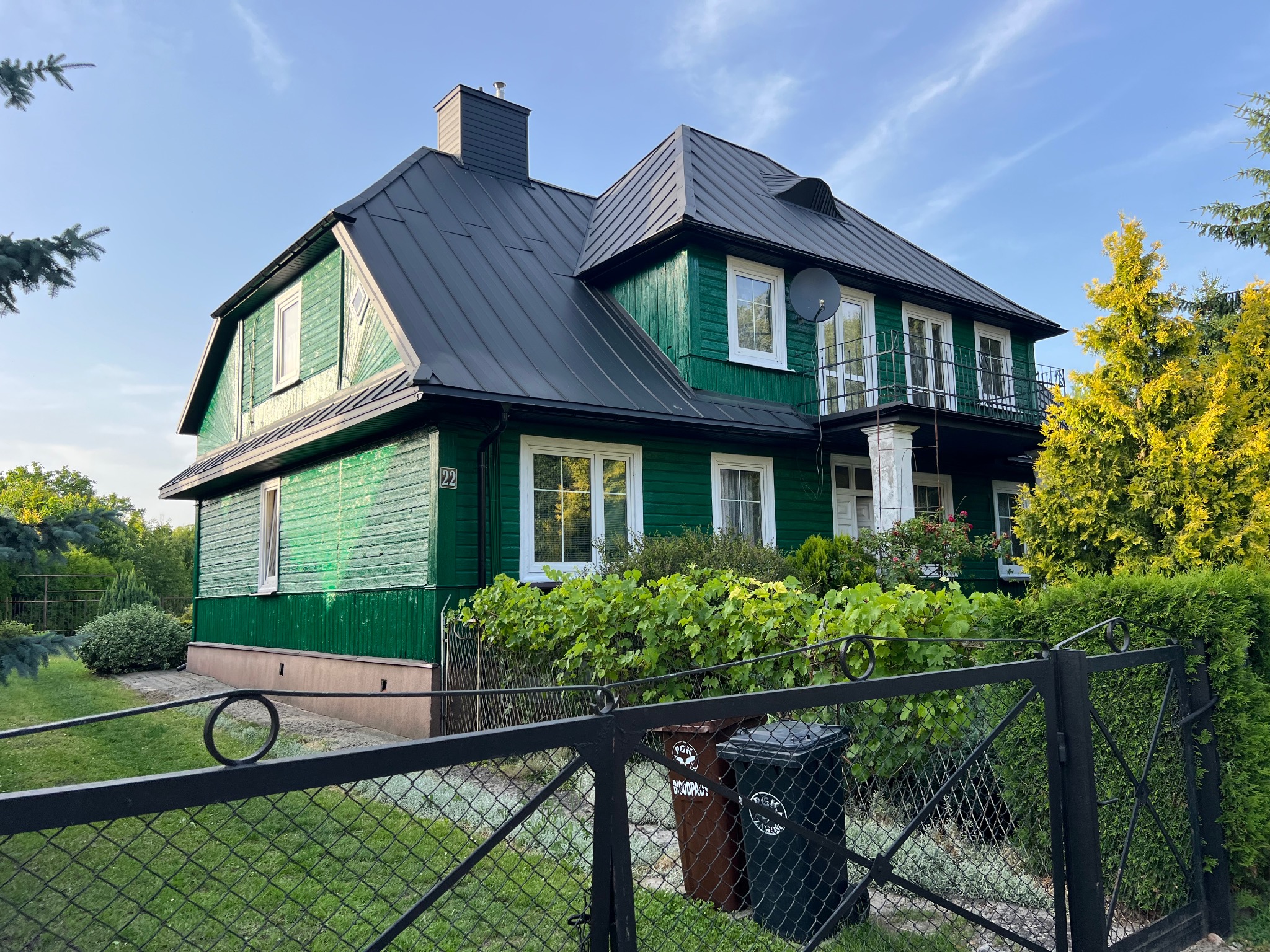 Dwupiętrowy dom z ciemnym dachem i zieloną elewacją, widoczny zza metalowego ogrodzenia, z numerem 22 na ścianie.