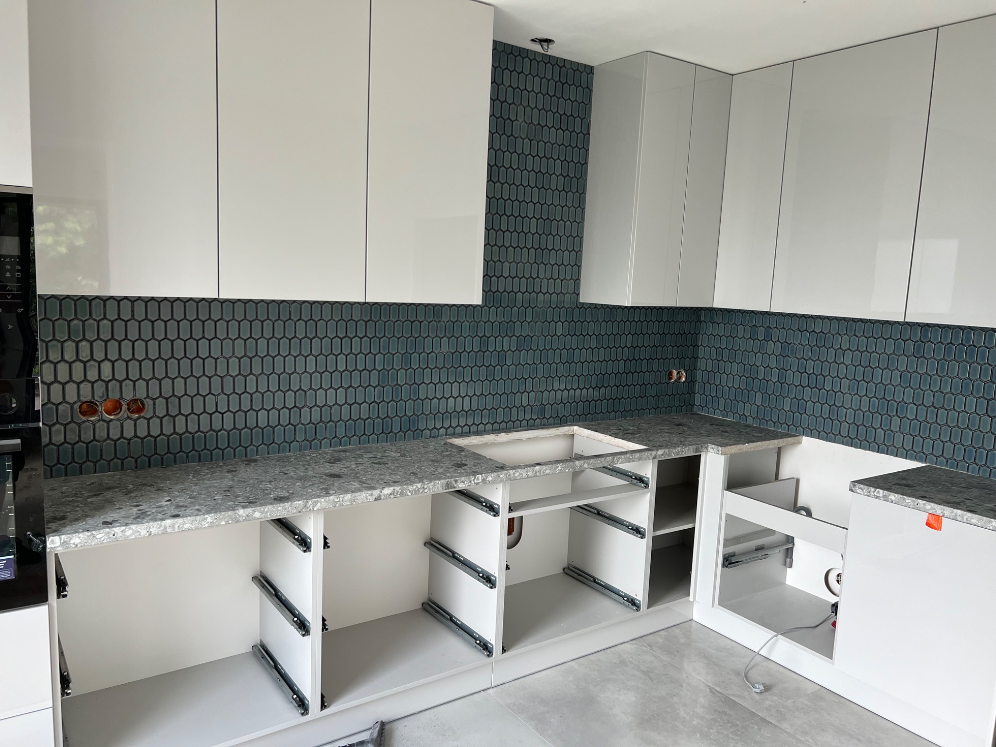 Montaż białych szafek kuchennych z szarym blatem i niebieską mozaiką w tle, widoczne szyny do szuflad, instalacja w Józefowie w trakcie realizacji.
