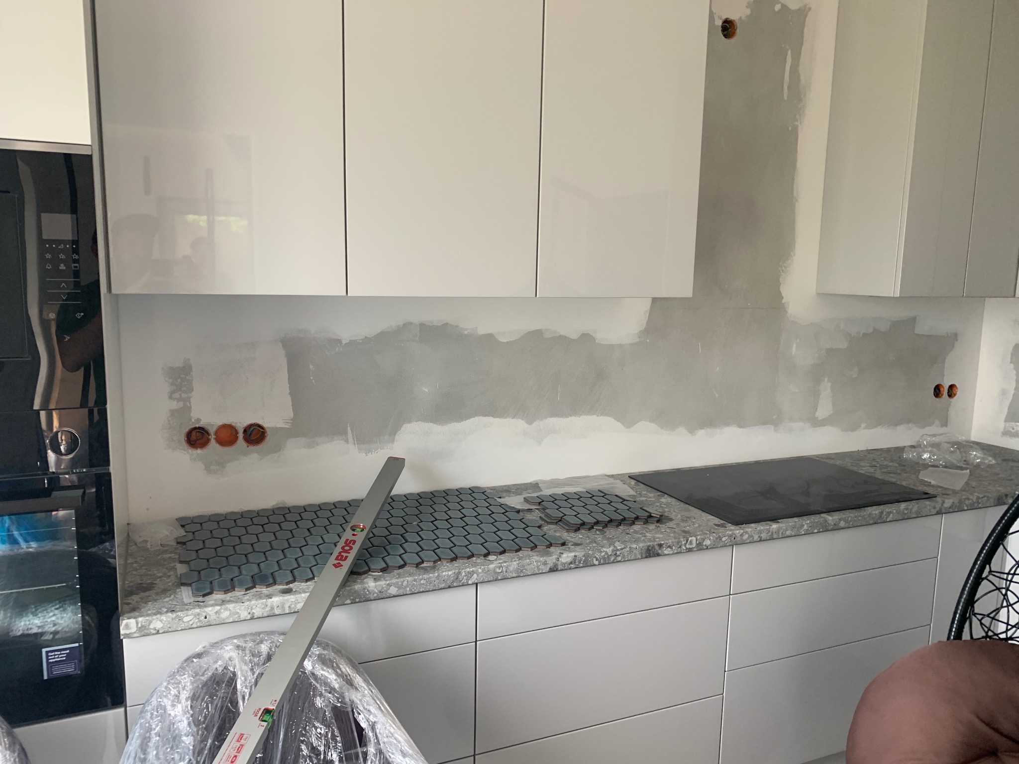 Przygotowanie do montażu mozaiki heksagonalnej w kuchni, widoczne surowe ściany nad blatem, poziomica oparta o blat, fragment płyty grzewczej, białe szafki kuchenne na górze i na dole.